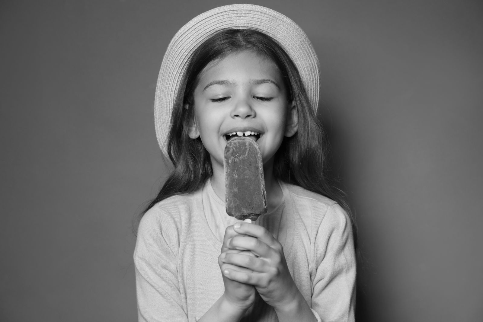 Black-and-white portrait of a smiling girl in a sun hat eating an ice lolly