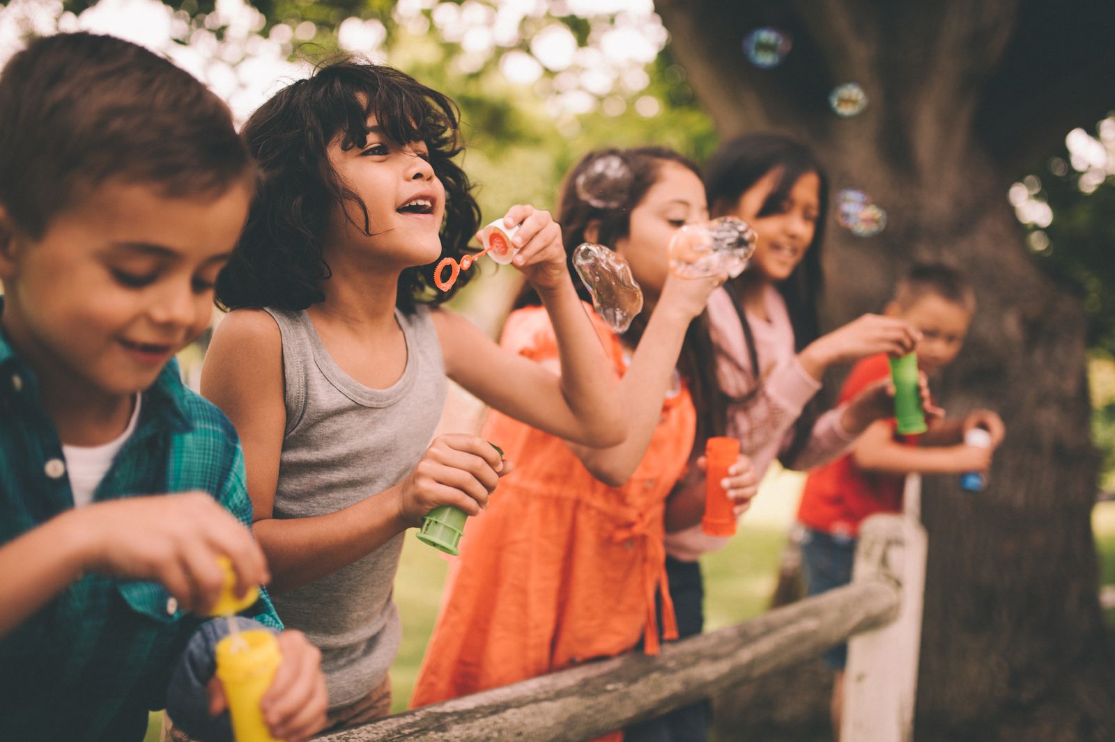 Children laughing and blowing soap bubbles outdoors by a wooden fence