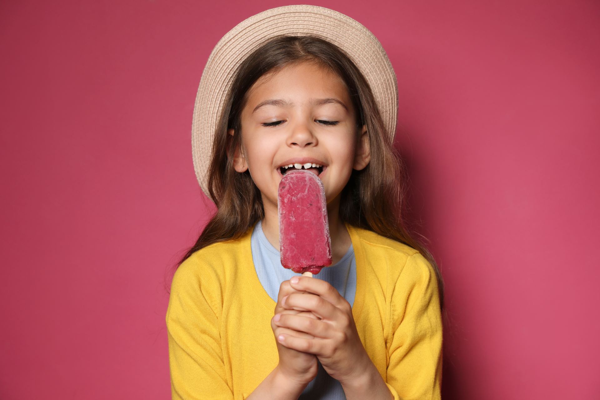 Smiling girl in a straw hat and yellow cardigan eating a pink ice lolly against a pink wall