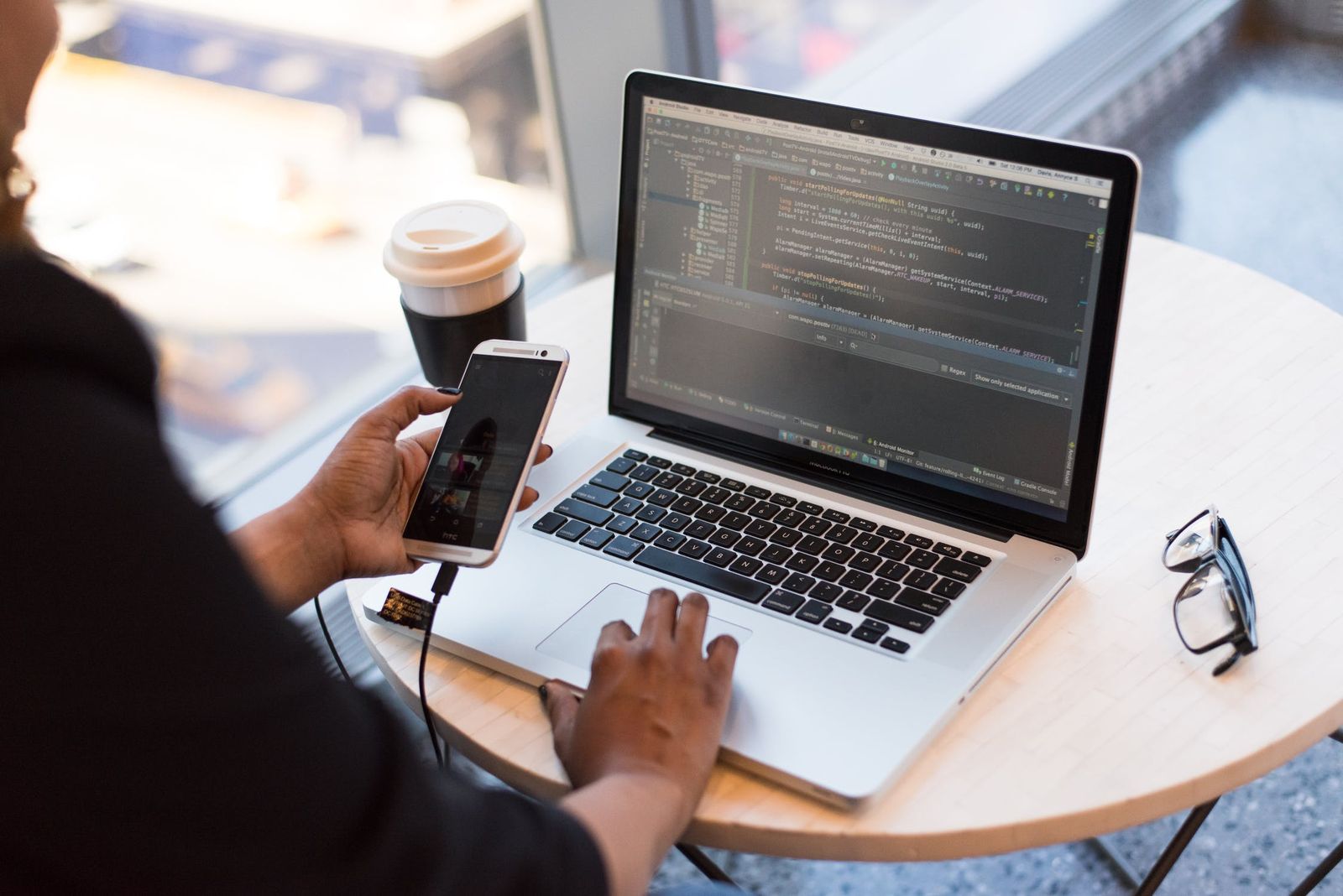 Hands holding a phone beside a laptop showing code, with a coffee cup on a café table