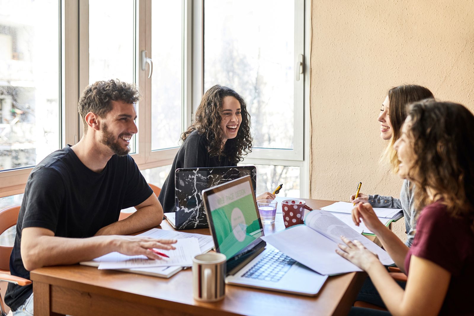Four students laughing while studying together with laptops and notes around a table