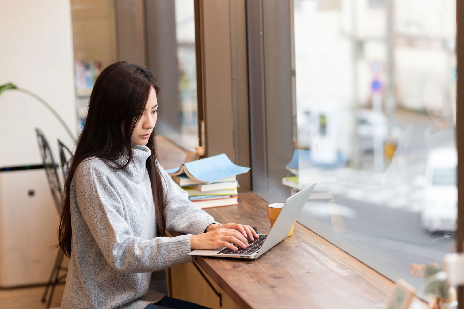 Student working on a laptop at a window counter beside a stack of books